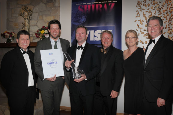 Five men and women, all in formal wear, pose for a photo. One man holds a crystal carafe trophy.