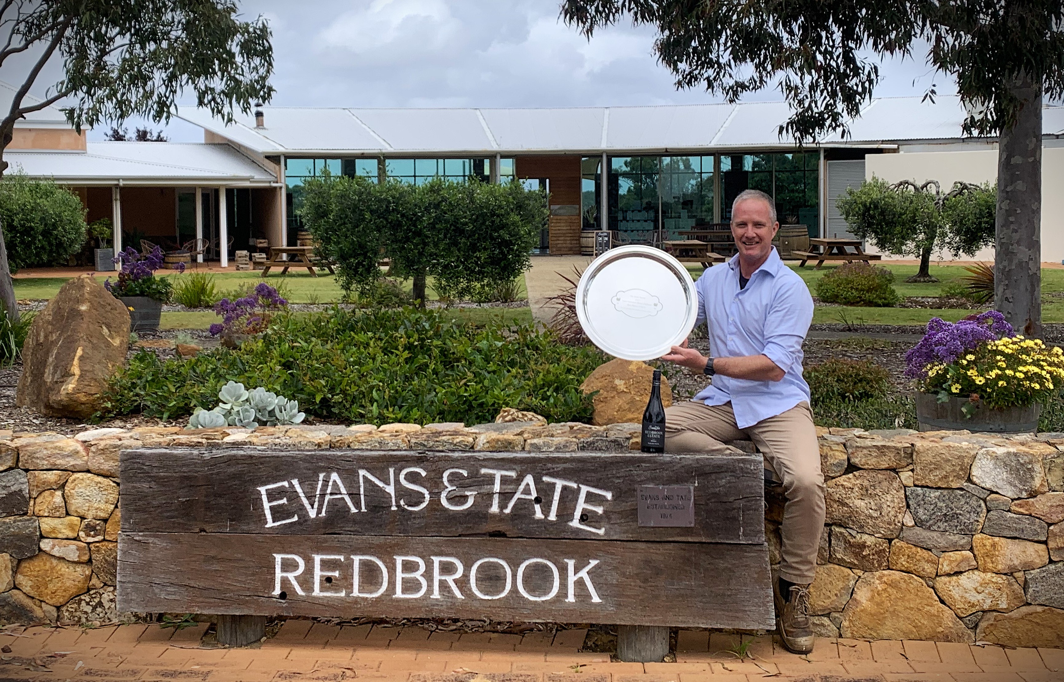 A man sits on a sandstone wall holding an engraved silver plate. He sits next to a large wooden sign for Evans & Tate Redbrook, established 1974. A bottle of red wine sits on the sign. Garden and winery buildings behind.