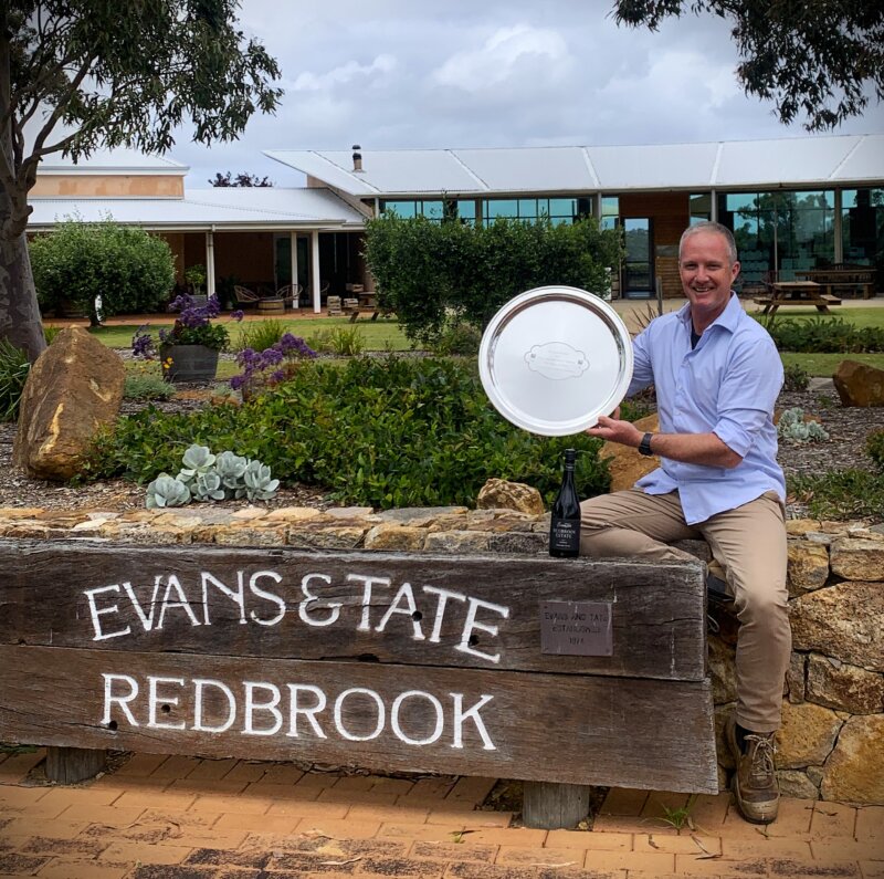 A man sits on a sandstone wall holding an engraved silver plate. He sits next to a large wooden sign for Evans & Tate Redbrook, established 1974. A bottle of red wine sits on the sign. Garden and winery buildings behind.